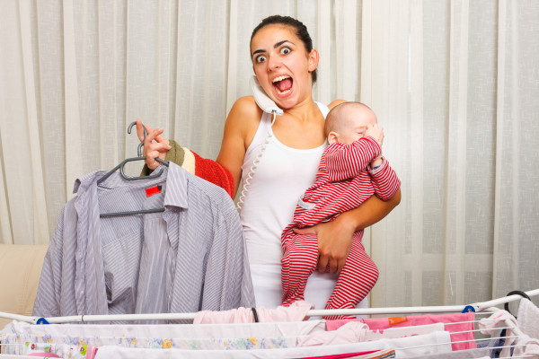 Angry young mother fed up with doing the laundry, holding her crying child and talking on the phone in the same time.