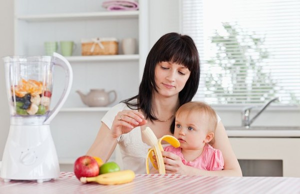 Attractive brunette woman pealing a banana while holding her baby on her knees in the kitchen