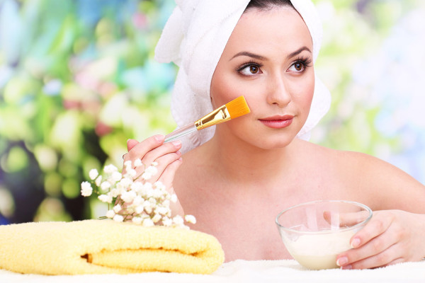 Beautiful young woman with cream for face mask towel on her head on bright background